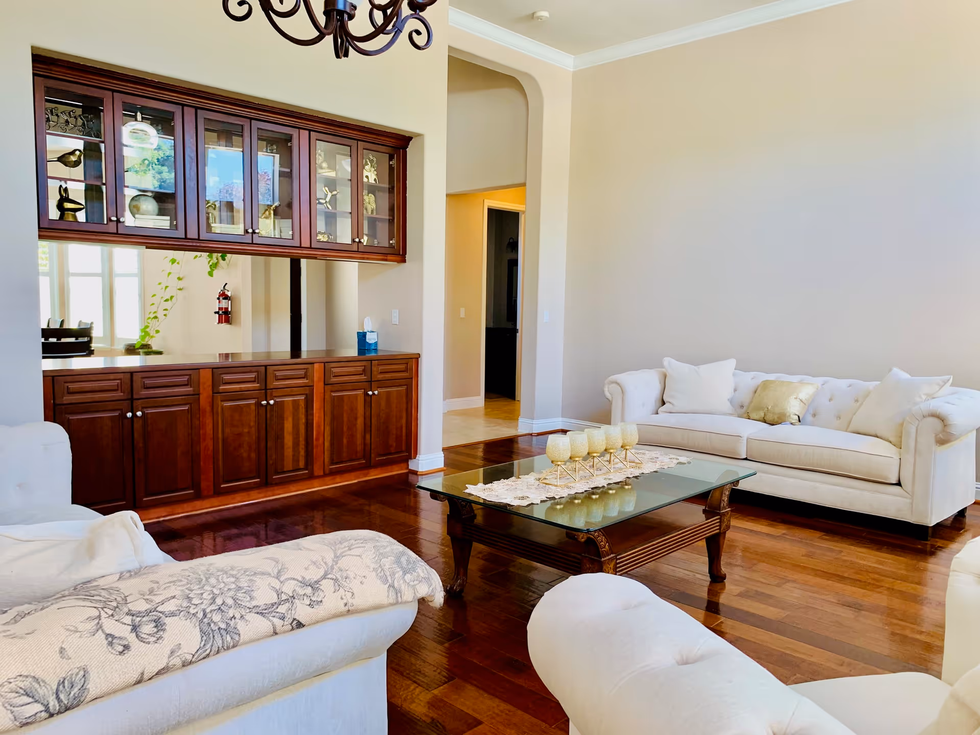 A bright living room with polished wooden floors, a glass coffee table with decorative candles, and cream-colored sofas with cushions. There is a wooden cabinet with glass doors mounted on the wall and a chandelier hanging from the ceiling. The walls are painted light beige.
