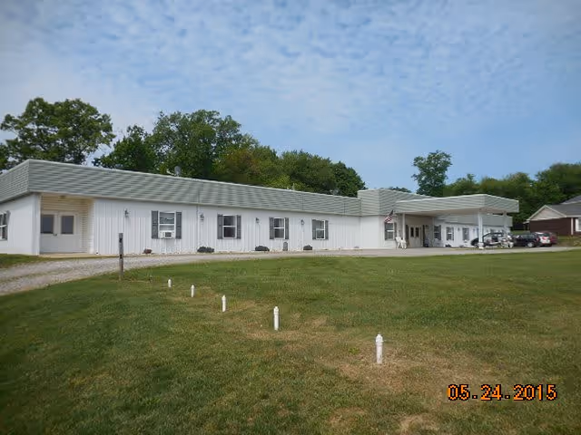 Single-story white building with a green roof, surrounded by grass and trees under a partly cloudy sky. The building has several windows and a covered entrance with an American flag. A few cars are parked near the entrance.