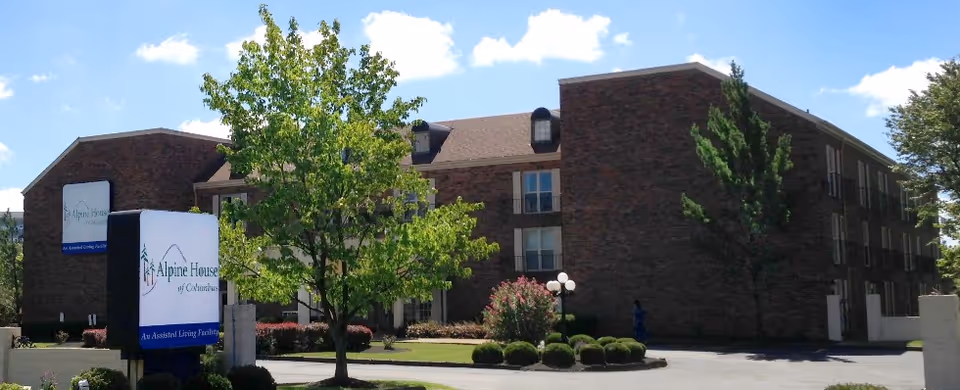 Exterior view of a brick senior living facility named Alpine House of Columbus with a sign in front, surrounded by trees and landscaping under a blue sky with some clouds.