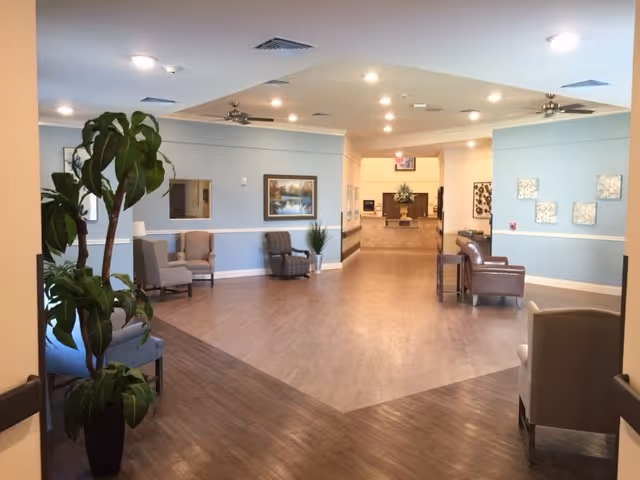 A spacious and well-lit interior hallway of a senior living facility with light blue walls and wood flooring. The hallway is furnished with several armchairs and small tables along the walls. There are decorative plants and framed artwork on the walls, with ceiling lights and fans providing illumination. The hallway leads to another room in the distance with more seating and floral arrangements.