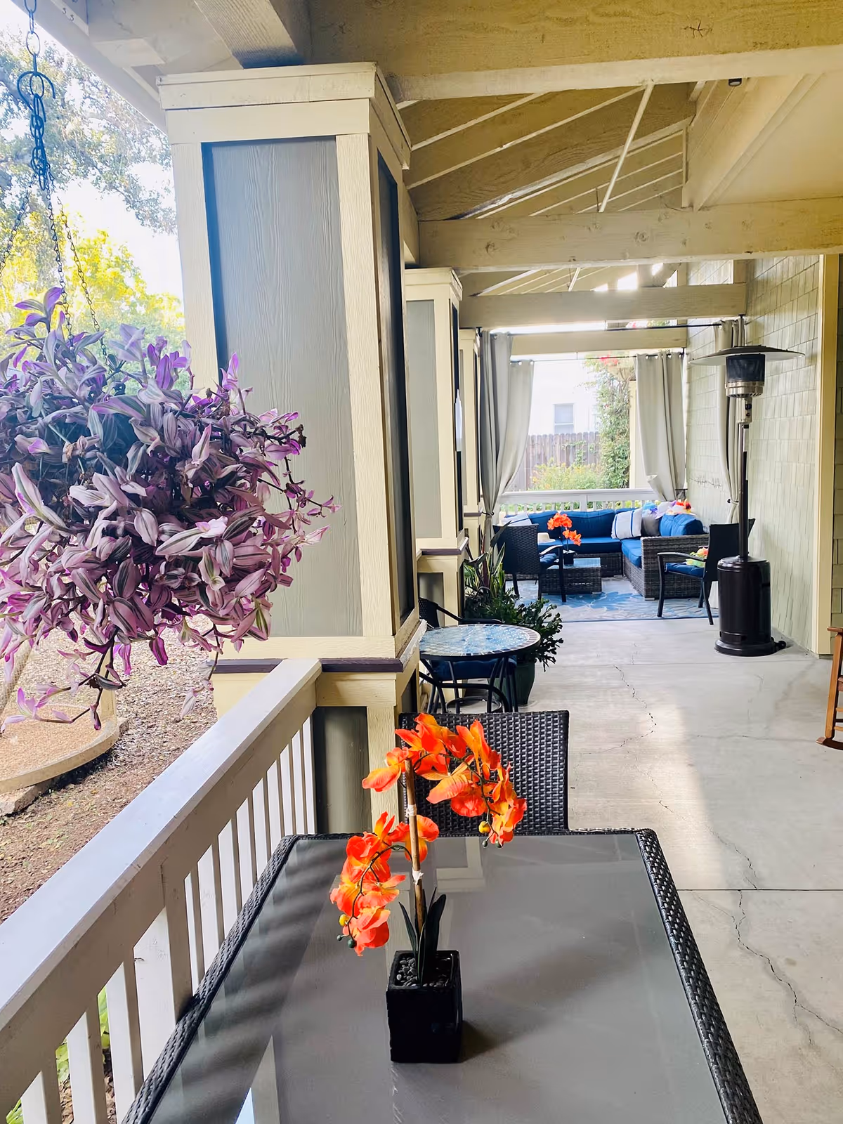 Covered outdoor patio area with a glass-top table and a black wicker chair in the foreground, decorated with a small potted plant with orange flowers. A hanging purple flower basket is visible on the left side. Further back, there is a seating area with blue cushioned wicker furniture, a small table, and a patio heater. The patio is shaded by a wooden roof with exposed beams, and light filters through the open sides.