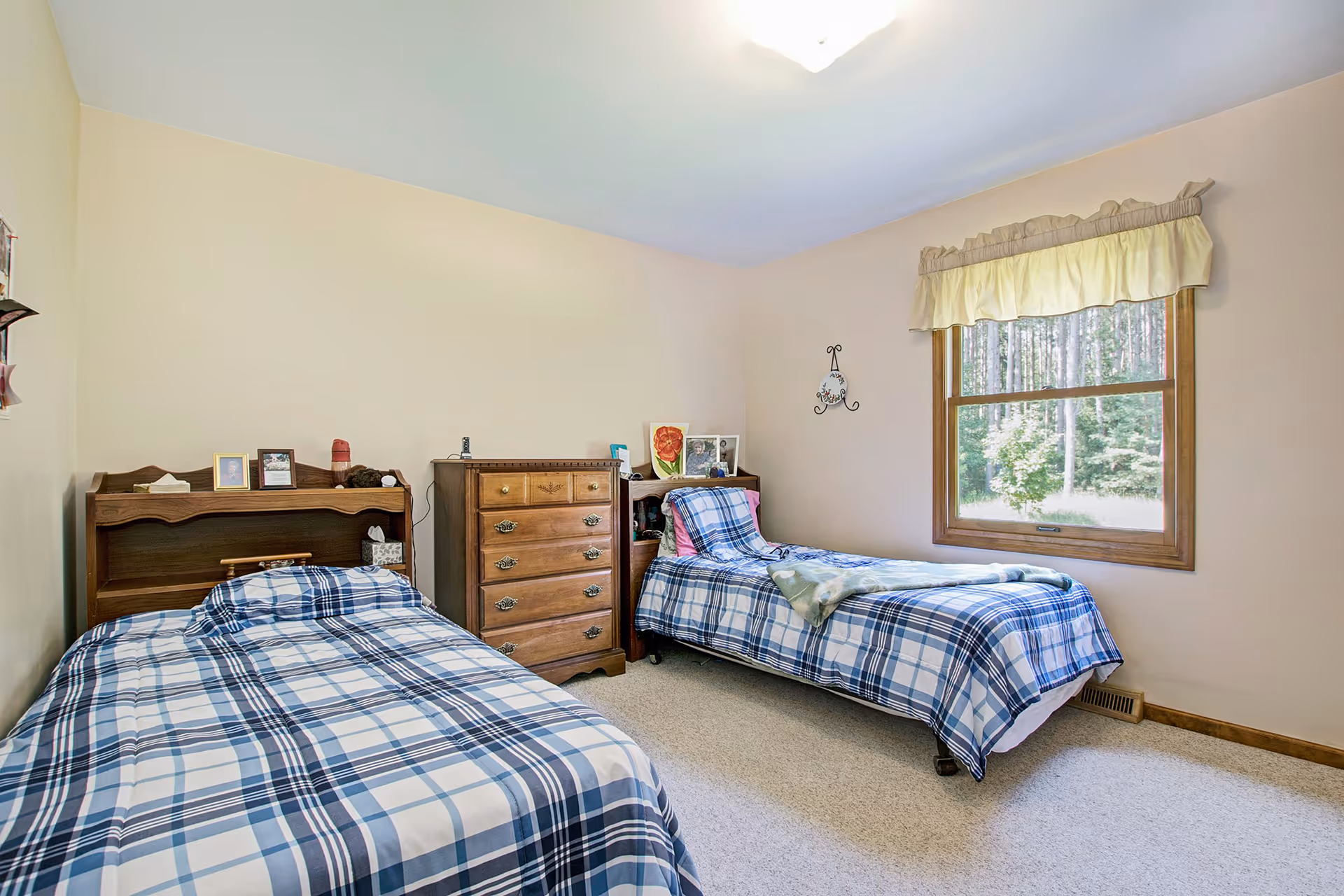 A simple bedroom with two twin beds covered in blue and white plaid bedding. Between the beds is a wooden chest of drawers and a small nightstand with framed photos and decorative items. A window with a yellow valance lets in natural light, showing a view of trees outside. The walls are painted light beige and the floor is carpeted.