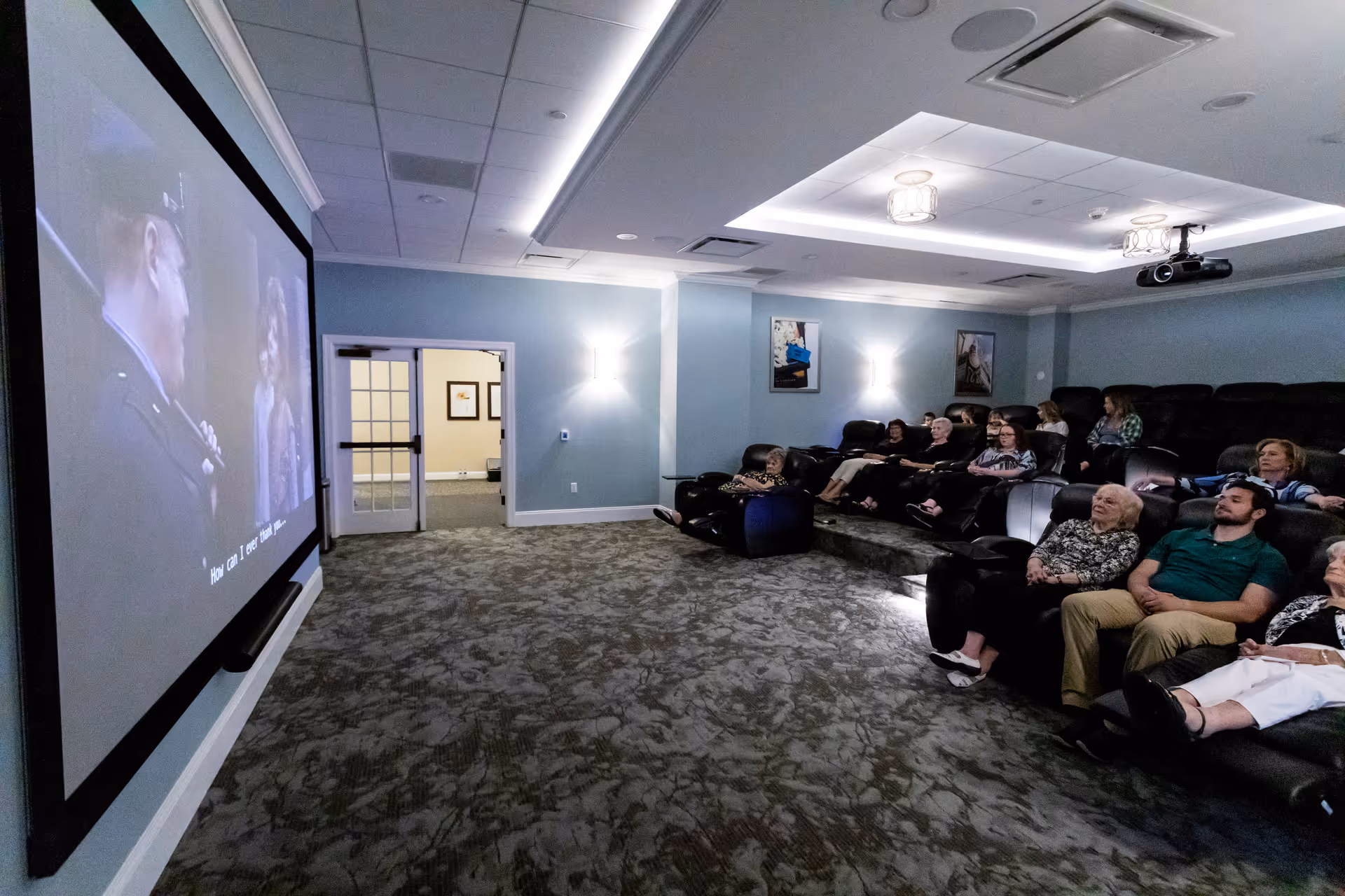 A group of people seated in a dimly lit movie theater room watching a film projected on a large screen. The room has tiered seating with black recliners and blue walls with framed pictures. Ceiling lights and wall sconces provide soft lighting.