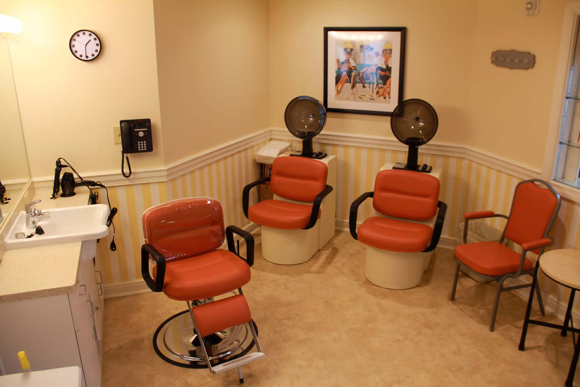 Interior of a salon area with three red salon chairs, two of which are positioned under hair dryers. There is a sink with a countertop and a wall-mounted phone on the left side. A framed picture hangs on the wall above the hair dryers, and there is a clock on the wall. The room has beige walls with a striped wallpaper border and a window on the right side.