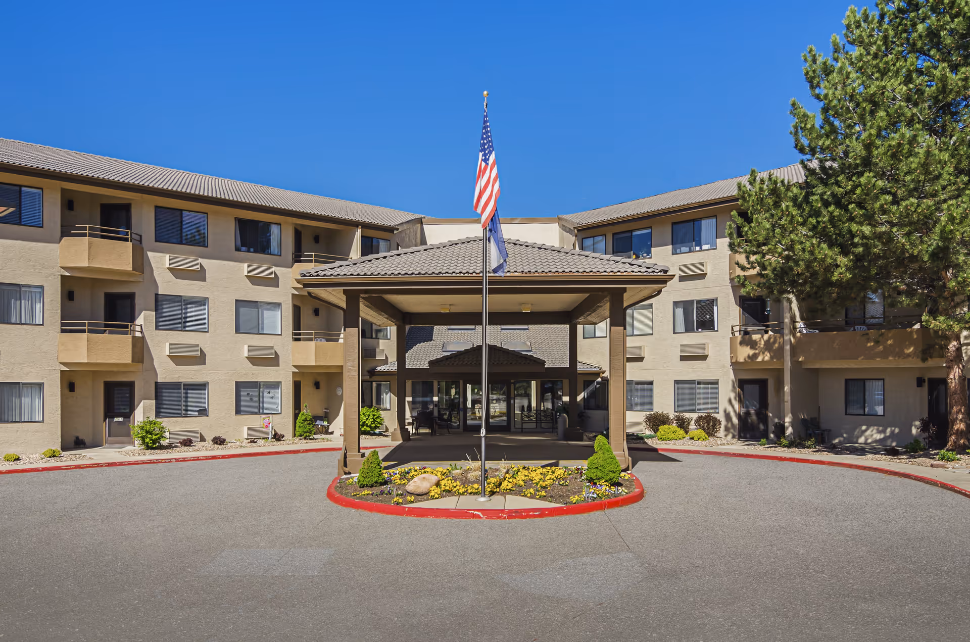 Front exterior view of a three-story senior living facility building with a covered entrance, an American flag on a flagpole, and landscaping with flowers and shrubs under a clear blue sky.