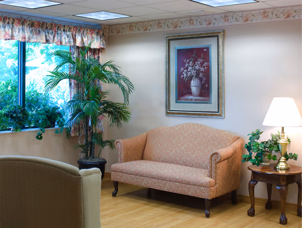 A cozy sitting area with a patterned peach-colored loveseat, a wooden side table with a brass lamp and green plants, a large potted palm plant near a window with floral curtains, and a framed floral painting on the wall.