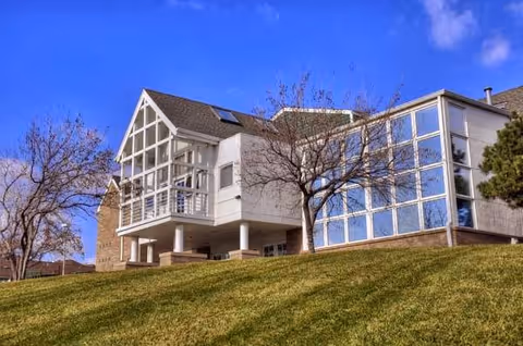 Exterior view of a modern assisted living facility building with large windows and a balcony, situated on a grassy hill with leafless trees under a clear blue sky.