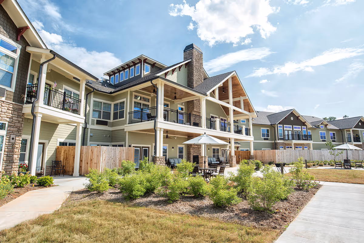 Exterior view of a two-story senior living building with balconies, patio seating, umbrellas, and a landscaped courtyard under a blue sky.
