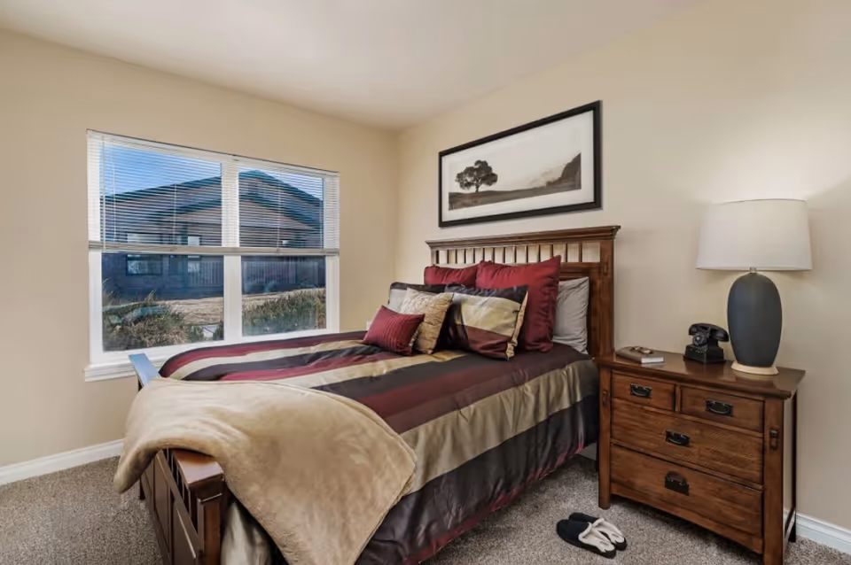 Bedroom with a wooden bed dressed in striped bedding and pillows, a nightstand with lamp and phone, and a window.