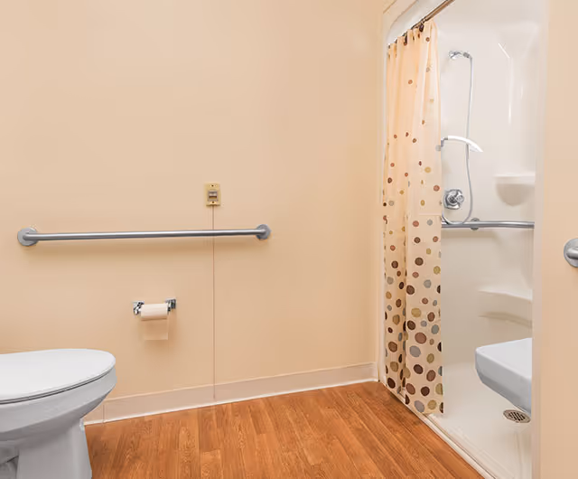 A bathroom with a toilet, a grab bar on the wall, a toilet paper holder, and a shower with a polka dot shower curtain. The floor has wood-like flooring and the walls are painted beige.