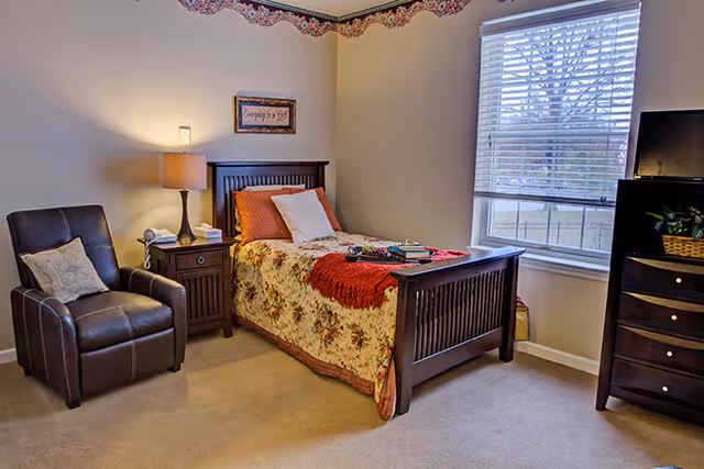 A cozy bedroom with a single wooden bed covered with a floral bedspread and a red throw blanket. Next to the bed is a wooden nightstand with a lamp, a phone, and a tissue box. A comfortable brown armchair with a decorative pillow is placed beside the nightstand. There is a window with white blinds letting in natural light, and a wooden dresser with a TV and a basket on top is positioned near the window. The walls are light-colored with a floral border near the ceiling.