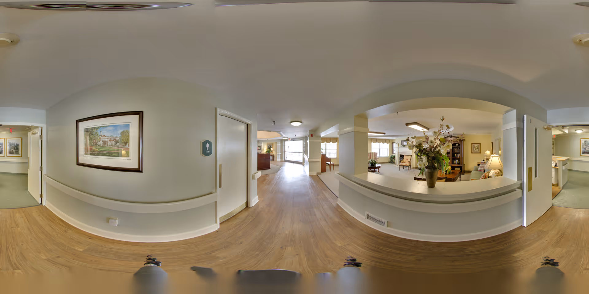 A wide-angle view of a senior living facility interior hallway with light wood flooring and cream-colored walls. To the right, there is a cozy living room area with armchairs, a lamp, a bookshelf, and a large floral arrangement on a ledge. The hallway leads to a reception desk in the distance with large windows letting in natural light. Several framed paintings hang on the walls.
