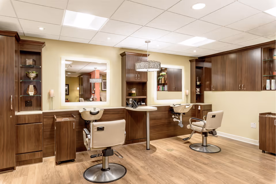 Interior view of a salon area in a senior living facility with two beige salon chairs in front of two large mirrors mounted on a wall with wooden cabinetry. The room has wooden flooring, soft yellow walls, and modern lighting fixtures including a decorative hanging lamp.
