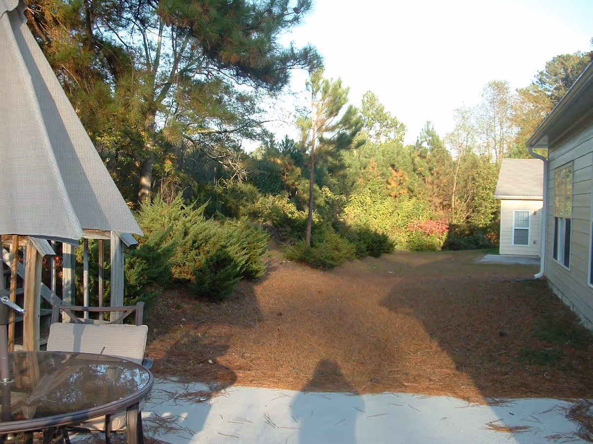 Outdoor patio area with a glass-top table and chairs under a large beige umbrella, surrounded by trees and bushes. A beige building is visible on the right side with windows and a small concrete patio.