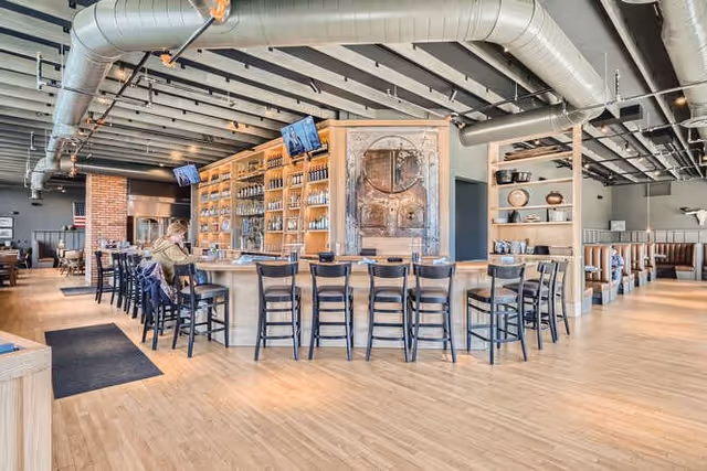 Interior view of a modern dining area with a large wooden bar counter surrounded by black chairs. Shelves behind the bar hold various glassware and bottles. The ceiling features exposed ductwork and beams, and there are multiple TV screens mounted above the bar. A person is seated at the bar, and there are additional tables and chairs in the background.