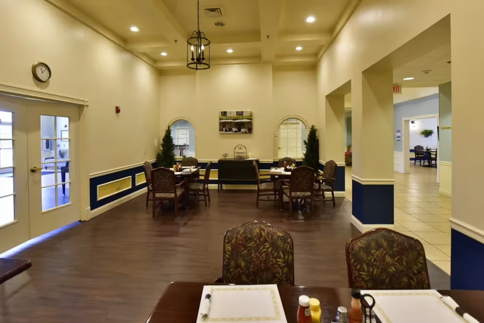 Interior view of a dining area in a senior living facility with multiple tables and chairs arranged for meals. The room has cream-colored walls with blue wainscoting, a wooden floor, and decorative plants near the back wall. There is a clock on the left wall above a set of double doors with glass panes, and a painting hangs on the back wall between two arched windows.