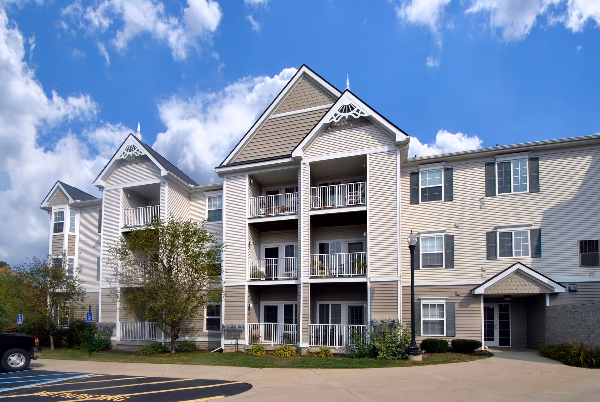 Exterior view of a three-story residential building with balconies, beige siding, white trim, and decorative gables under a partly cloudy blue sky. There are trees and shrubs in front of the building and a parking area with marked spaces, including handicapped spots.