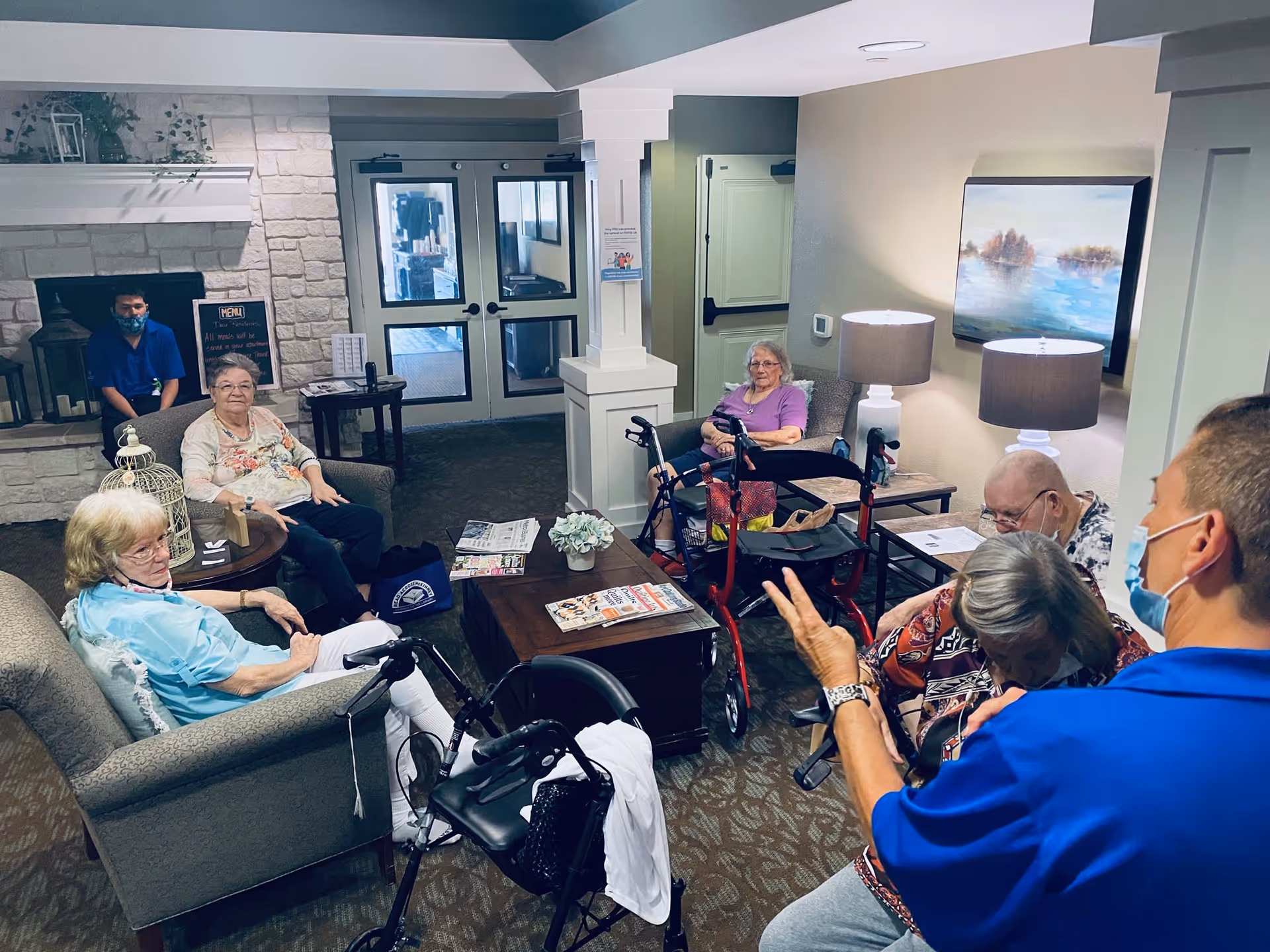 A group of elderly people sitting and interacting in a cozy common area with armchairs, a coffee table with magazines, and a fireplace. Two staff members wearing blue shirts and face masks are assisting the residents. The room is warmly lit with table lamps and decorated with a painting on the wall.