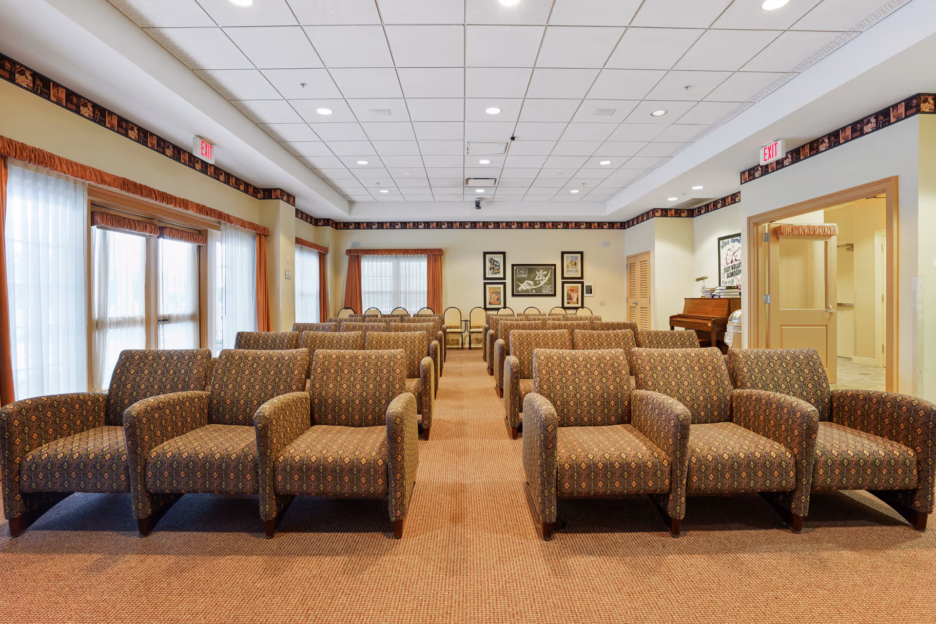 A well-lit room with multiple rows of patterned armchairs arranged facing forward, large windows with sheer curtains on the left side, framed pictures on the far wall, and a piano on the right side near an open doorway.
