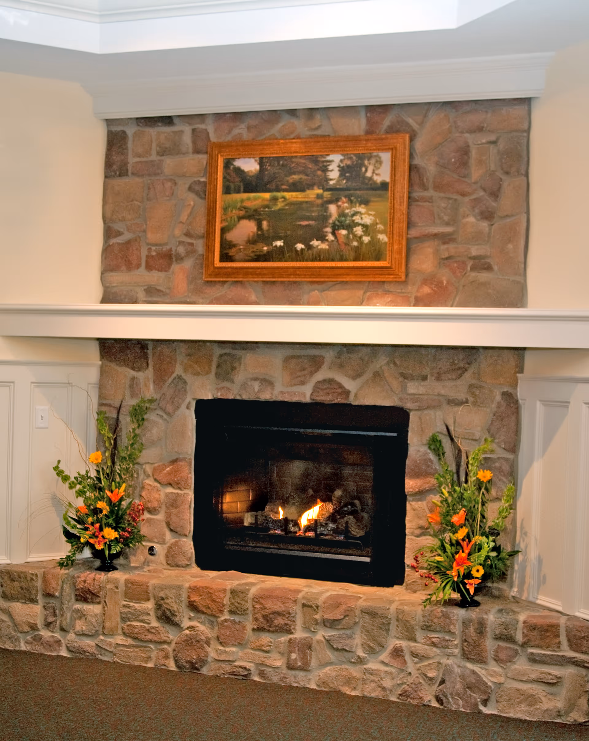 A cozy stone fireplace with a fire burning inside, flanked by two floral arrangements with orange and yellow flowers. Above the fireplace is a framed painting depicting a serene outdoor scene with a pond and greenery.