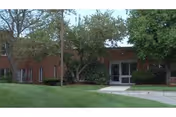 Exterior view of a brick building with large windows and a glass entrance door, surrounded by green grass, trees, and shrubs.