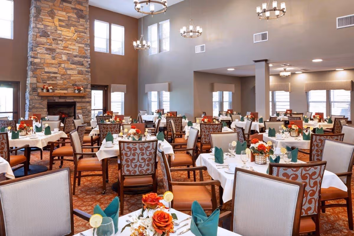 A spacious dining room with multiple tables set for a meal. Each table is covered with a white tablecloth, decorated with floral centerpieces, and has green folded napkins. The room features a high ceiling with large windows letting in natural light, a stone fireplace on one side, and elegant chandeliers hanging from the ceiling.