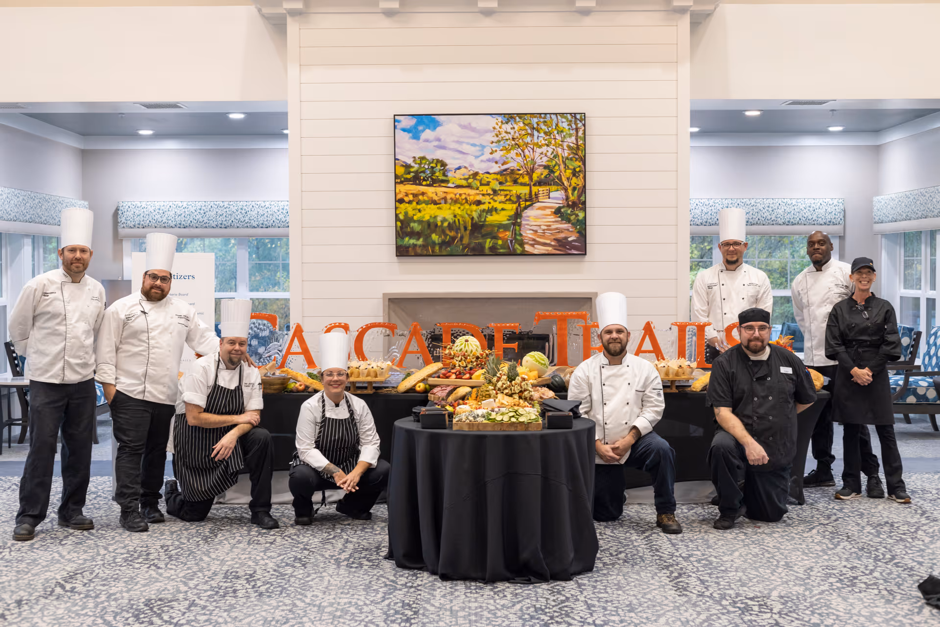 A group of chefs and kitchen staff posing in a dining area behind a table filled with an assortment of food and decorative items. The background features a white shiplap wall with a colorful landscape painting and large letters spelling out part of 'Cascade Trails'.