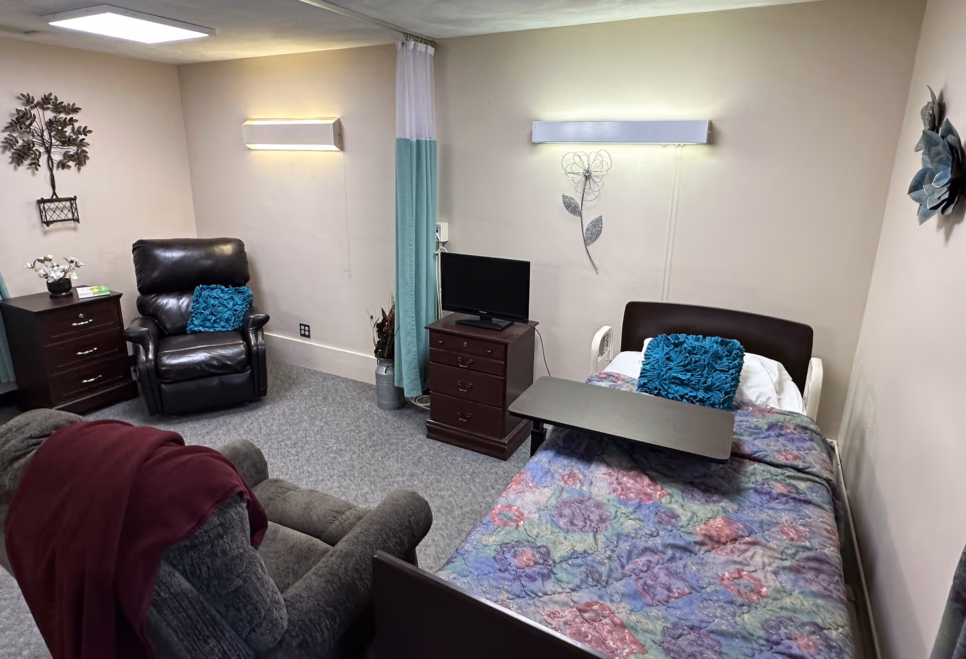 A furnished senior care bedroom with a single bed and overbed table, two recliner chairs, a TV on a dresser, and wall decorations.