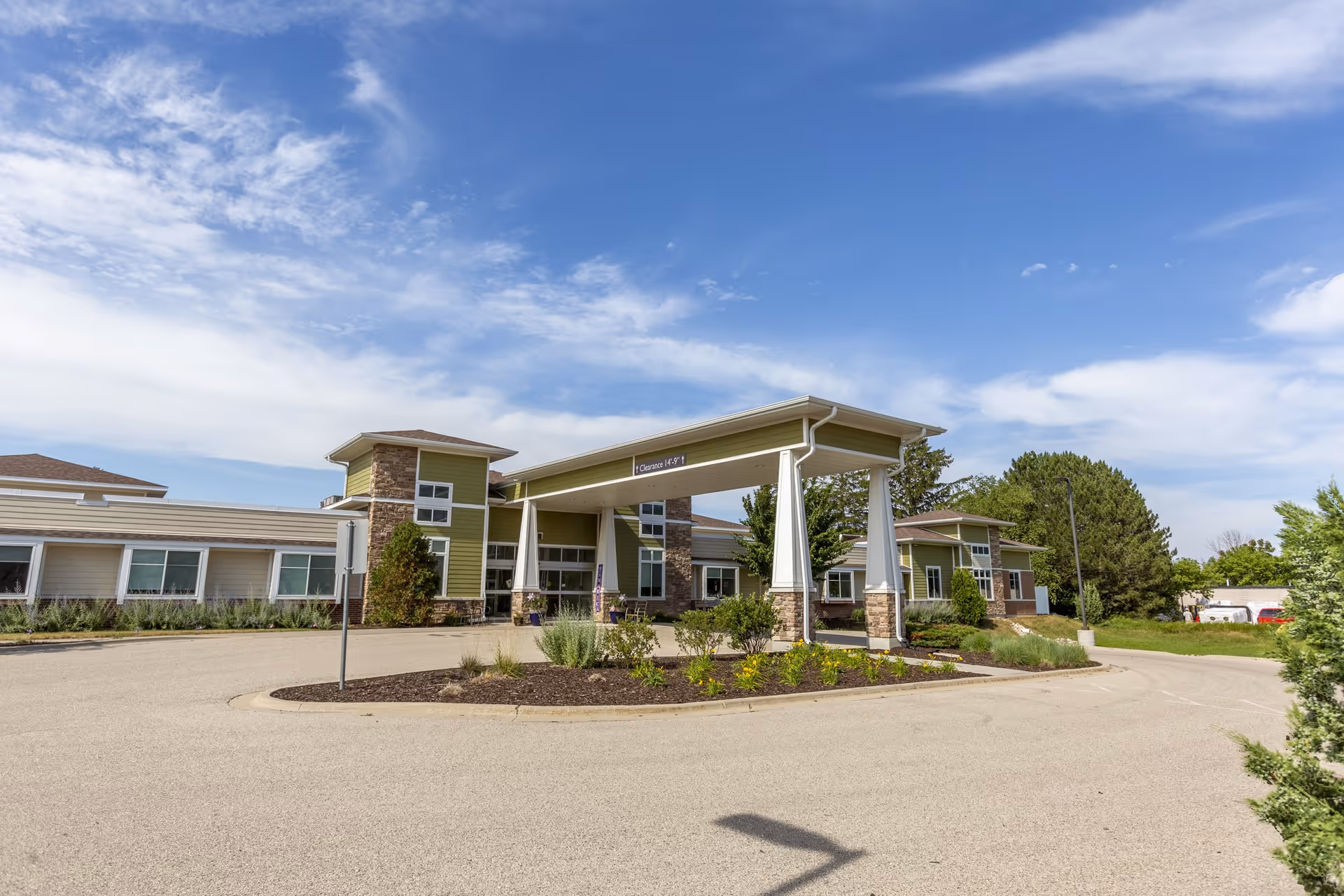 Exterior view of StoryPoint Mequon senior living facility showing a single-story building with green and beige siding, stone accents, large windows, and a covered entrance with white pillars under a partly cloudy blue sky.