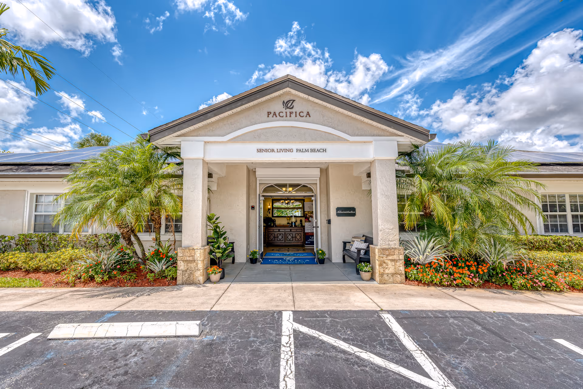 Front entrance of the Pacifica senior living facility with a covered portico, palm landscaping, and parking under a blue sky.