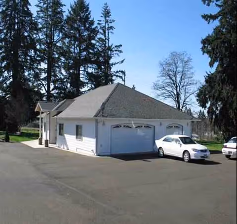 White single-story building with a two-car garage and cars in a driveway, tall trees behind.