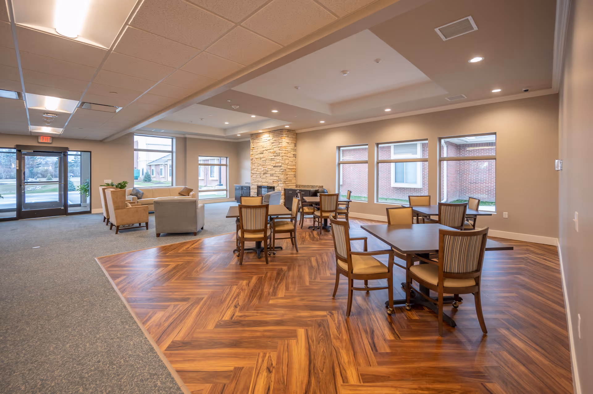 Spacious senior living common area with wood-patterned flooring, tables and chairs, lounge seating, and large windows.