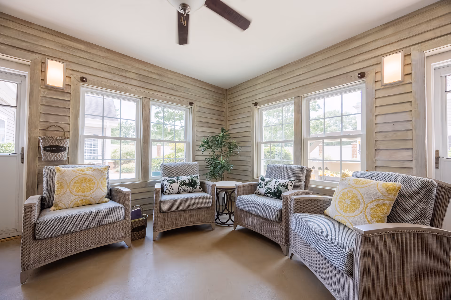 A cozy indoor seating area with four wicker armchairs arranged around a small round table. The chairs have cushions with yellow and green patterned pillows. The room has large windows on two walls, allowing natural light to fill the space. The walls are covered with light-colored wooden paneling, and there is a ceiling fan above.