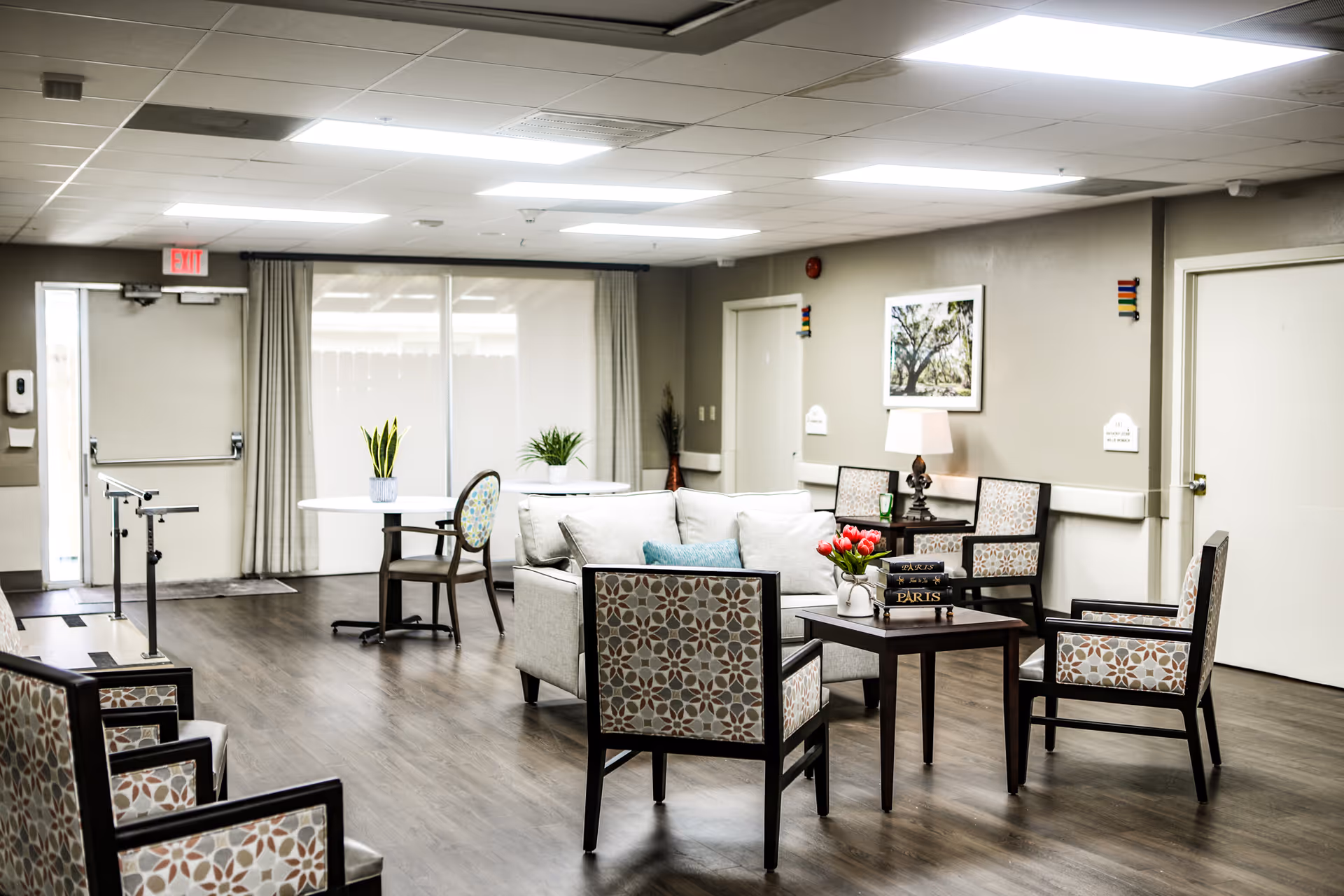 A bright and spacious living room area in a senior living facility with patterned armchairs arranged around a small wooden table holding a vase of red tulips and two books titled 'Paris'. A white sofa with cushions is positioned near the chairs. In the background, there is a round table with a plant, a lamp on a side table, framed artwork on the wall, and large windows covered with curtains letting in natural light. The floor is wooden, and the ceiling has recessed lighting.