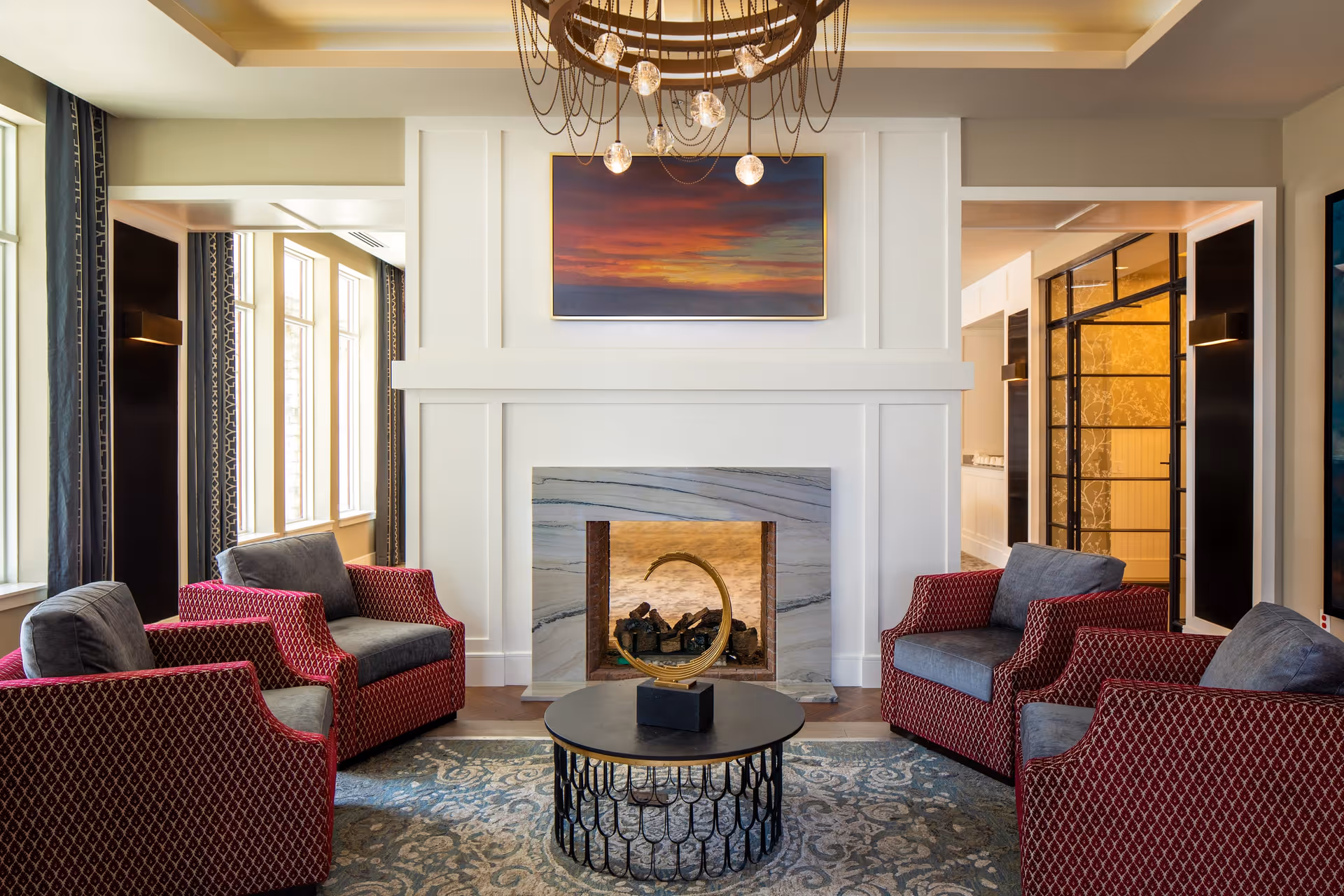 Elegant seating area with four patterned armchairs around a round coffee table facing a decorative fireplace and chandelier.