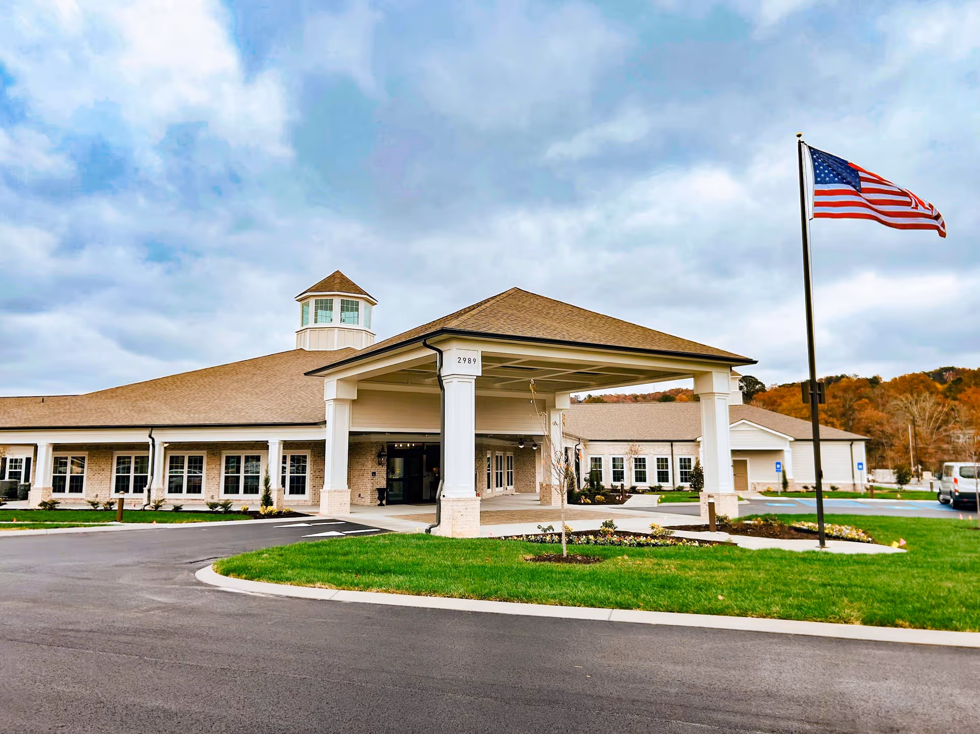 Exterior view of a senior living facility building with a covered entrance, multiple windows, and a cupola on the roof. There is a large American flag on a flagpole to the right, a paved driveway, and landscaped grass and flower beds in front of the building under a cloudy sky.