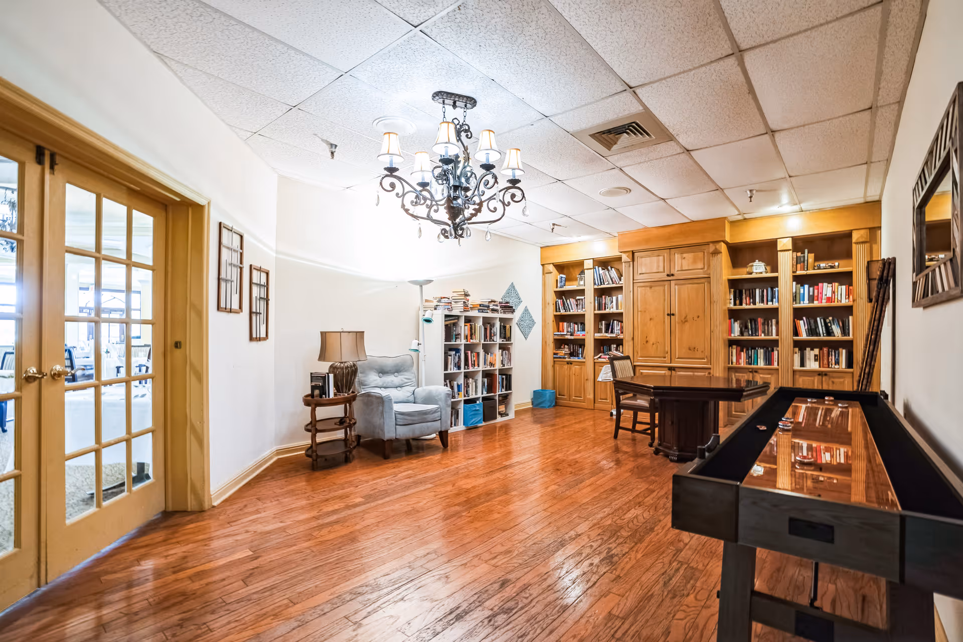 A cozy interior room with wooden flooring, a chandelier hanging from the ceiling, a comfortable armchair next to a small side table with a lamp, bookshelves filled with books, a wooden table with chairs, and a shuffleboard table. French doors with glass panes lead to another room.