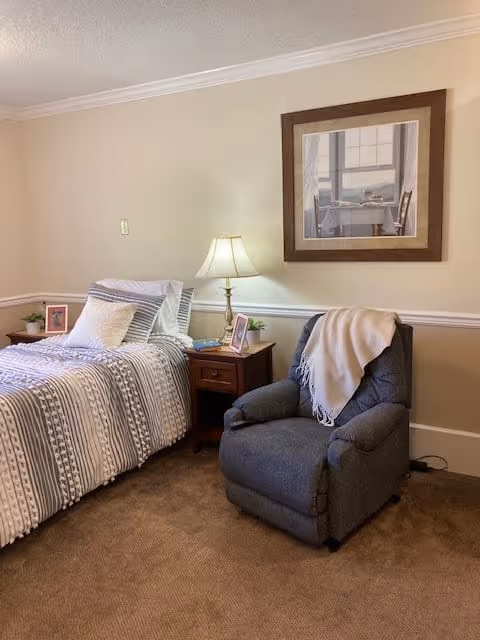 A cozy bedroom with a single bed dressed in striped bedding and multiple pillows. Next to the bed is a wooden nightstand with a table lamp, framed photos, and a small plant. A comfortable gray recliner chair with a light-colored throw blanket draped over the back is positioned nearby. A framed picture hangs on the beige wall above the chair. The room has beige carpet and white trim along the walls.