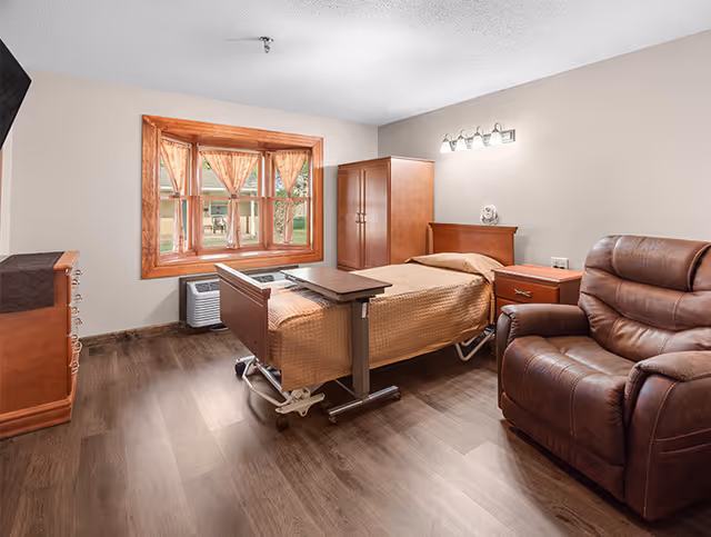 A senior living facility bedroom with a single bed covered in a brown quilt, a wooden nightstand with a lamp, a wooden wardrobe, a brown leather recliner chair, a wooden dresser, and a window with brown curtains letting in natural light.