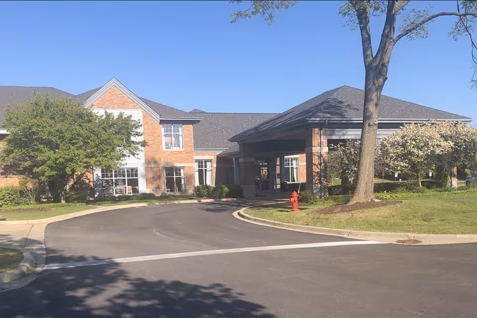 Front entrance of a brick senior living facility with a covered porte-cochere, curved driveway, trees, and a red fire hydrant.
