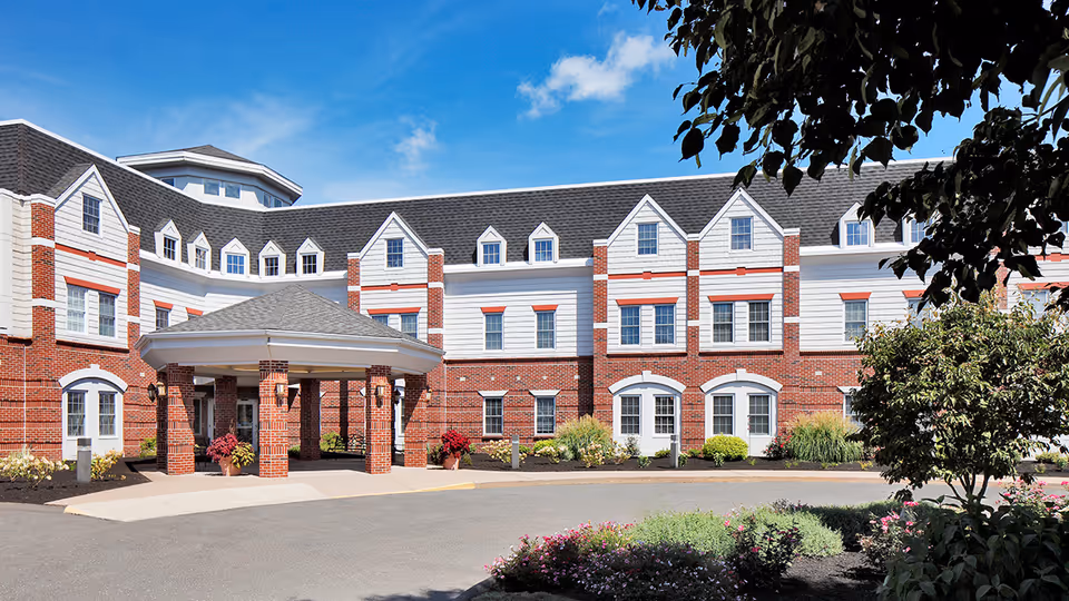 Front exterior view of Addison Place at Glastonbury, a multi-story brick and white siding building with a covered entrance, surrounded by landscaped bushes and flowers under a blue sky.