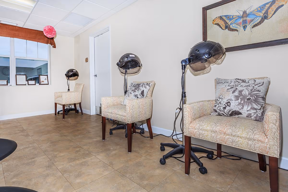 Interior view of a hair salon area in an assisted living facility with three beige patterned chairs, each paired with a black hair dryer hood on a stand. The room has beige tiled flooring, cream-colored walls, a window with a brown valance and a pink decorative flower, and a framed butterfly artwork on the wall.