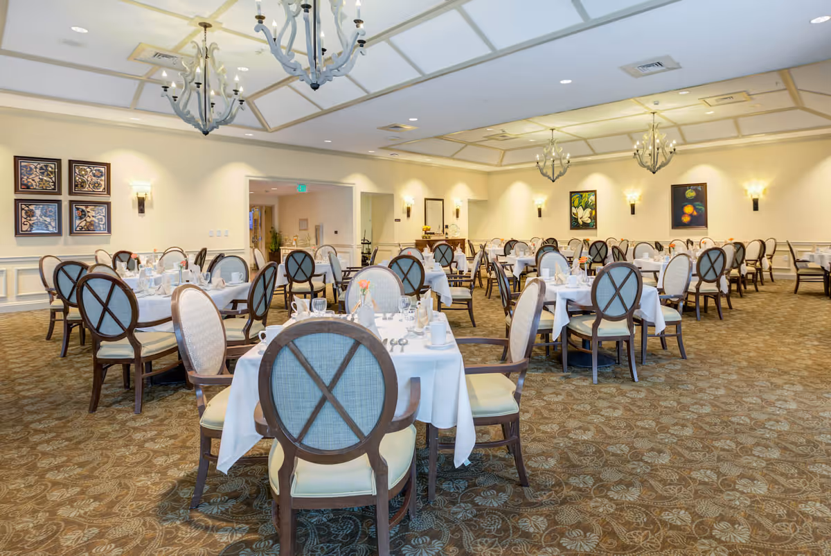 A spacious dining room with multiple round tables covered with white tablecloths, each set with plates, glasses, and silverware. The chairs have wooden frames with light-colored cushioned seats and backs. The room features patterned carpet, soft yellow walls with framed artwork, wall sconces, and elegant chandeliers hanging from the ceiling.