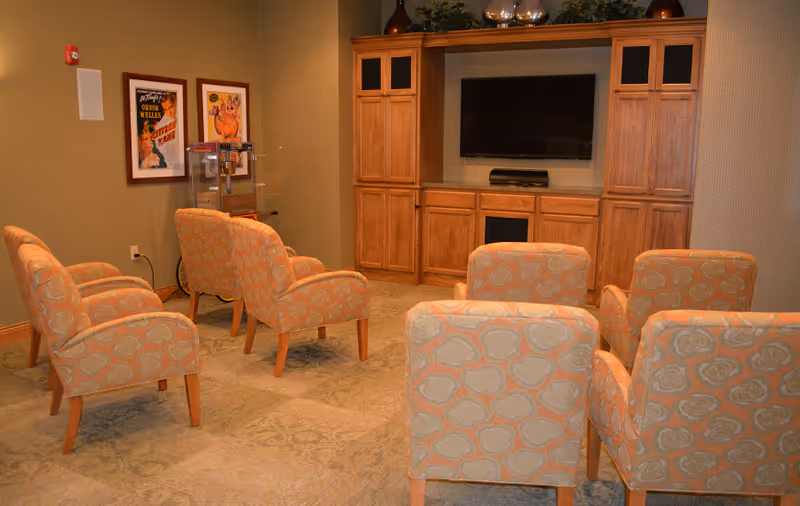 A cozy common area with six patterned armchairs arranged in two rows facing a large flat-screen TV mounted on a wooden entertainment center with cabinets. The room has beige walls, carpeted floor, framed vintage movie posters on the wall, and a popcorn machine in the corner.