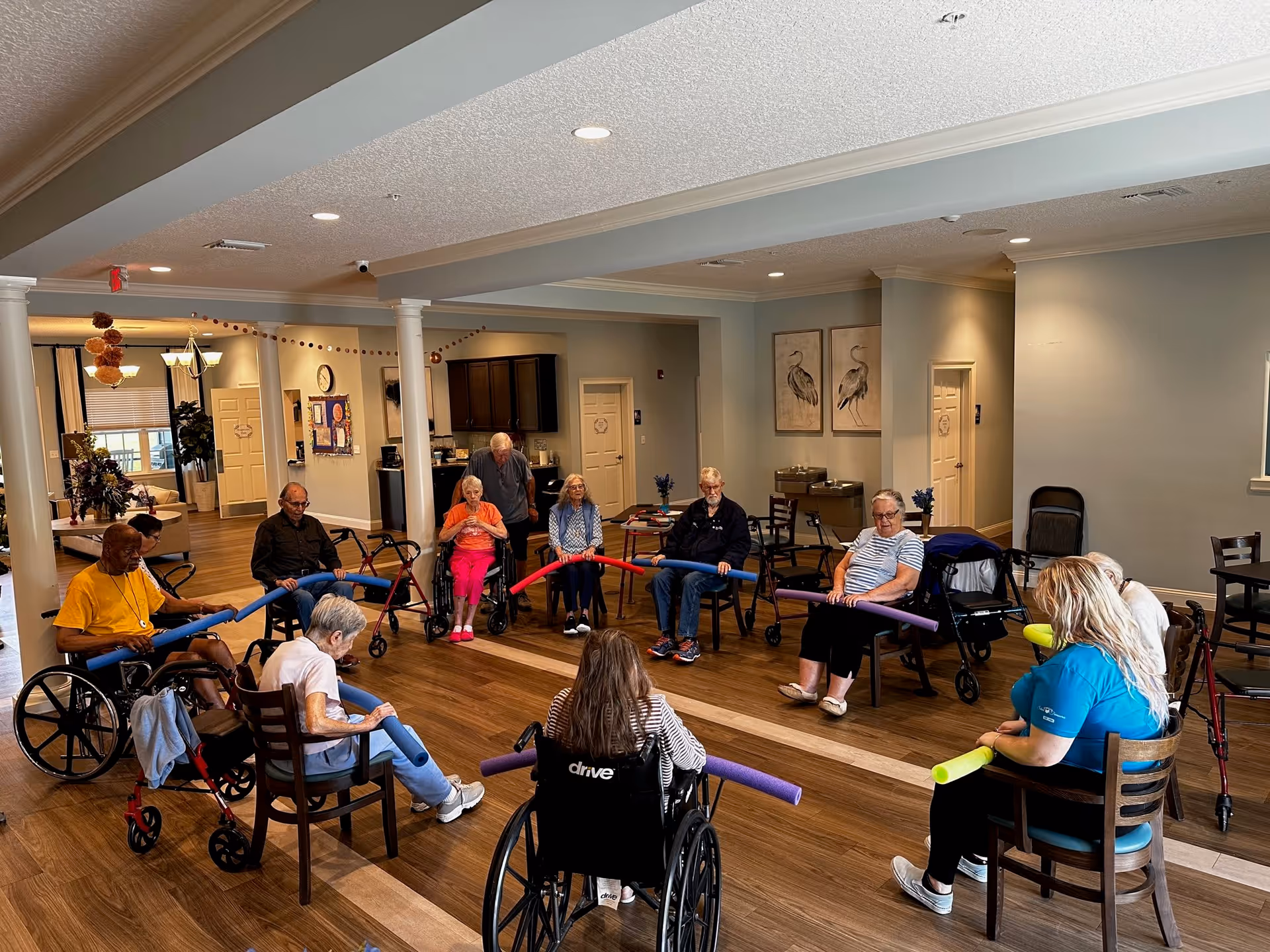 A group of elderly individuals seated in a circle in a spacious room, participating in a group activity using colorful foam noodles. Some participants are in wheelchairs, and a staff member is also present, engaging with the group. The room has wooden flooring, neutral-colored walls, and framed artwork on the walls.
