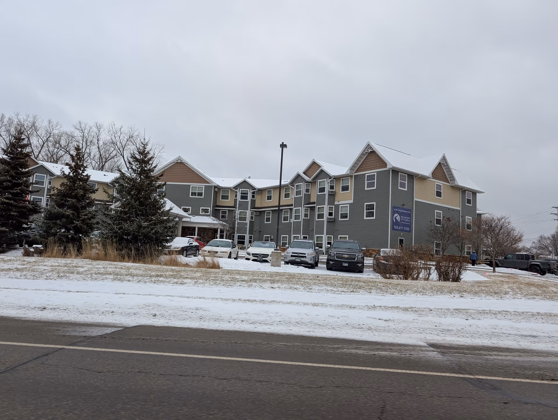 Front exterior of a multi-story residential building with parked cars and a light snow-covered lawn.