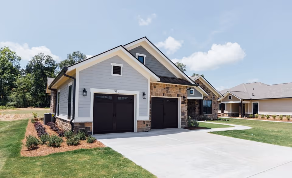 Front exterior of a single-story residential building with two dark garage doors, driveway, and landscaped lawn.