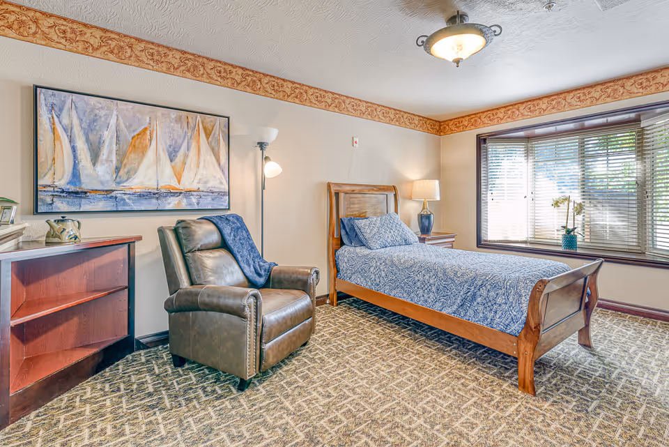A cozy bedroom featuring a wooden single bed with blue patterned bedding, a brown leather armchair with a blue throw draped over it, a wooden nightstand with a lamp, a large window with blinds and a potted orchid, a floor lamp, a wooden bookshelf, and a large painting of sailboats on the wall.