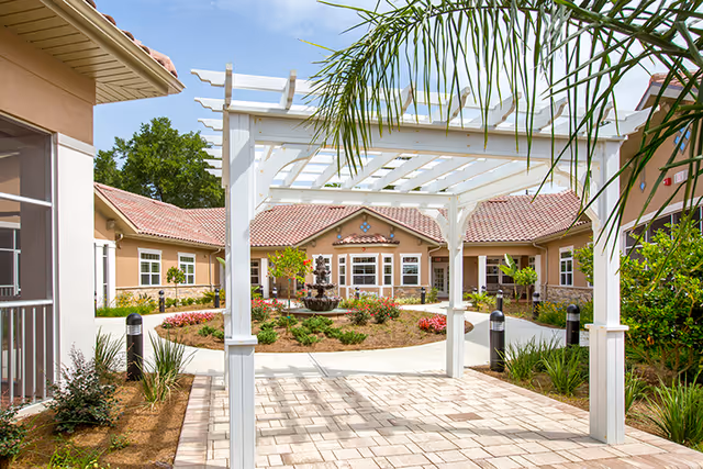 Outdoor courtyard area of Mission Oaks Assisted Living and Memory Care featuring a white pergola, paved walkway, landscaped garden with shrubs and flowers, a central water fountain, and beige buildings with red-tiled roofs in the background under a clear blue sky.