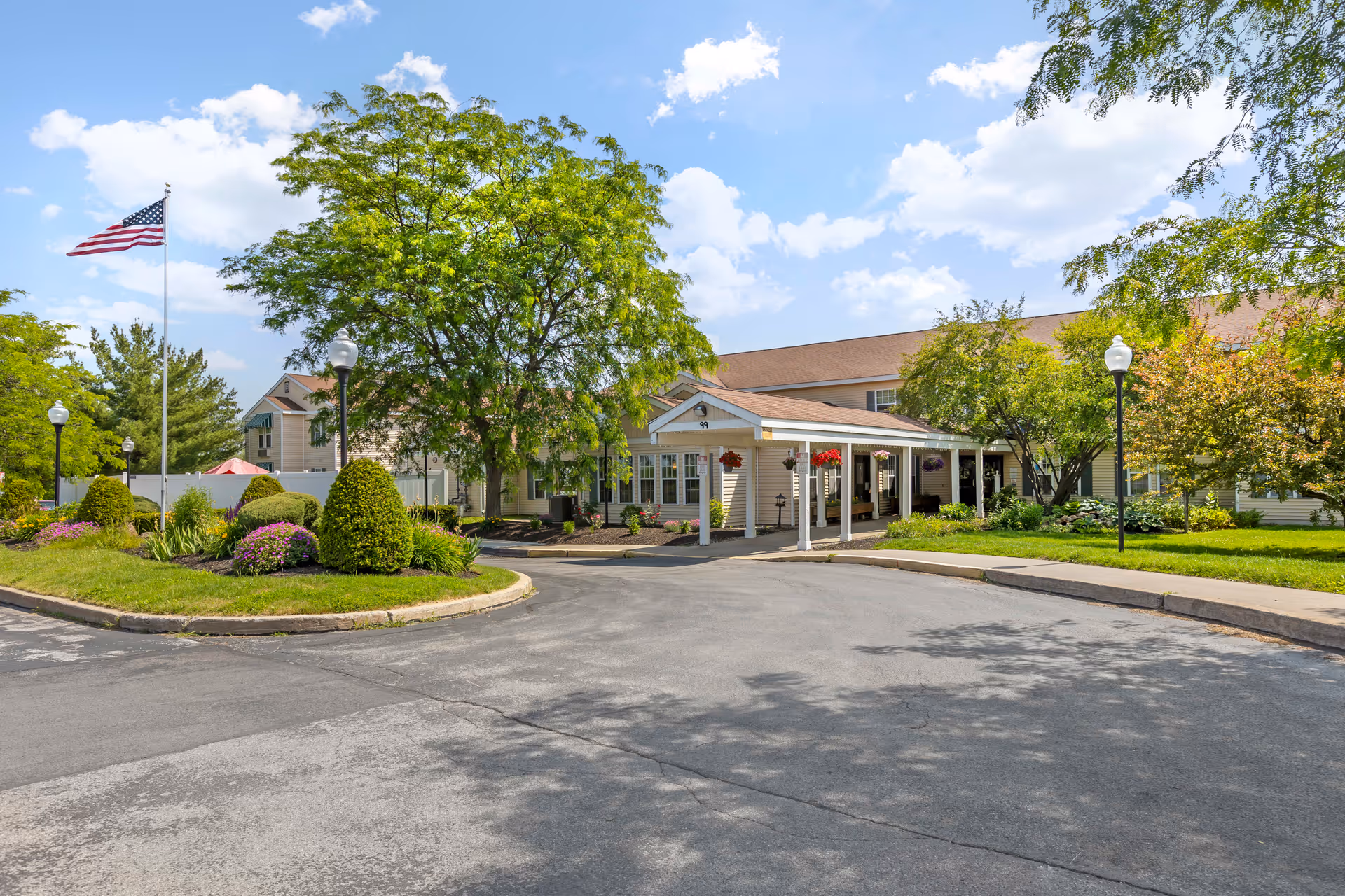 Exterior view of Brookdale Clinton senior living facility showing a driveway leading to the entrance with a covered porch. The building is surrounded by well-maintained landscaping including trees, bushes, and flowers. An American flag is flying on a flagpole to the left under a partly cloudy sky.